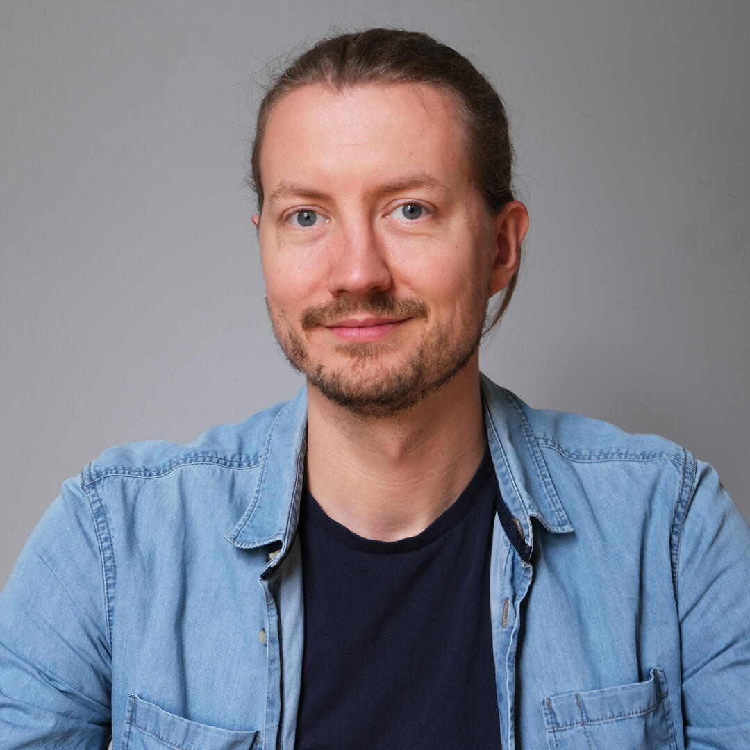 Photo d'un homme souriant aux cheveux longs attachés. Il porte une barbe courte et une chemise en jeans ouverte sur un t-shirt bleu.